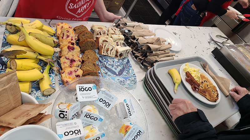 Breakfast foods arranged on a table, including bananas, sliced bread, muffins, packaged yogurt, and hot plates with prepared meals, as people serve food from the buffet setup.