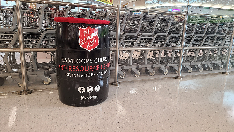 A black Salvation Army donation barrel labeled ‘Kamloops Church and Resource Centre’ placed inside a grocery store, with shopping carts lined up in the background.