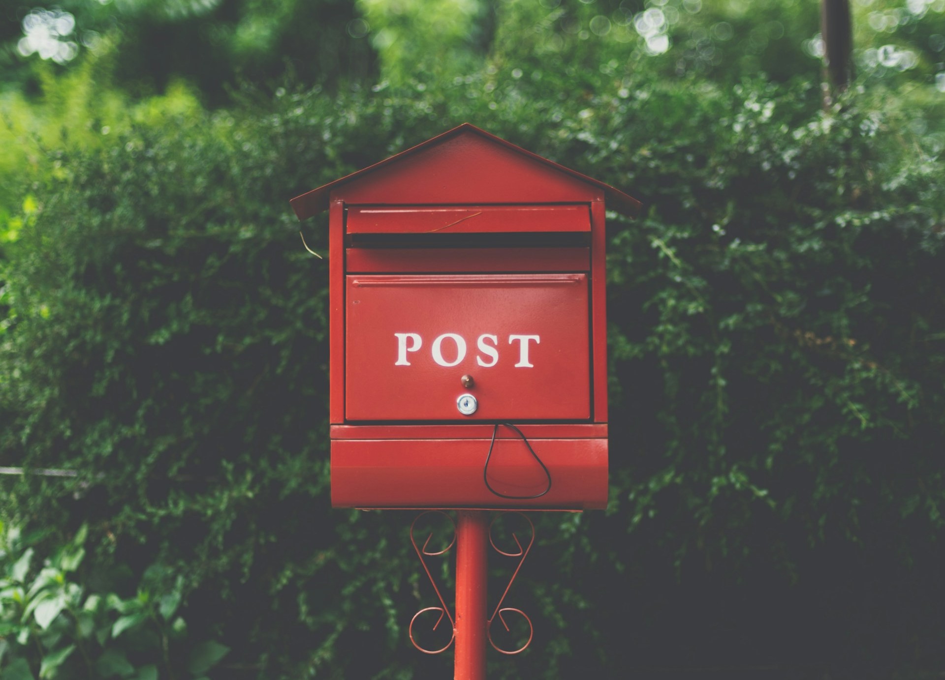 A red metal mailbox labeled ‘POST’ mounted on a post outdoors, set against a backdrop of green foliage.