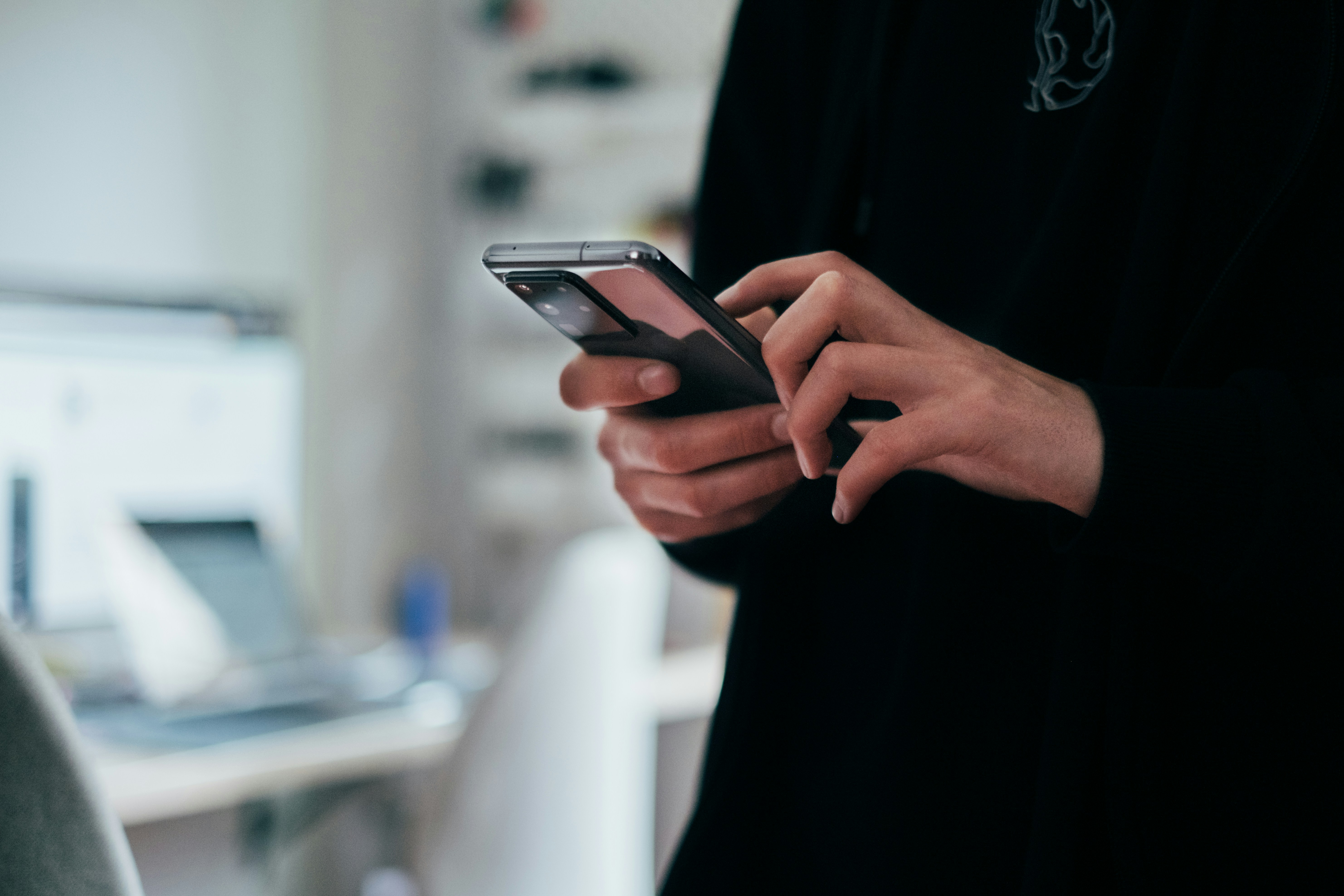 Close-up of hands holding and using a smartphone indoors, with a desk and laptop visible in the background.