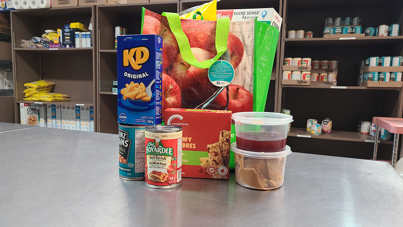 Food items arranged on a countertop, including boxed macaroni and cheese, canned goods, pasta, a bag of produce, and clear containers of prepared food, with pantry shelves stocked with supplies in the background.