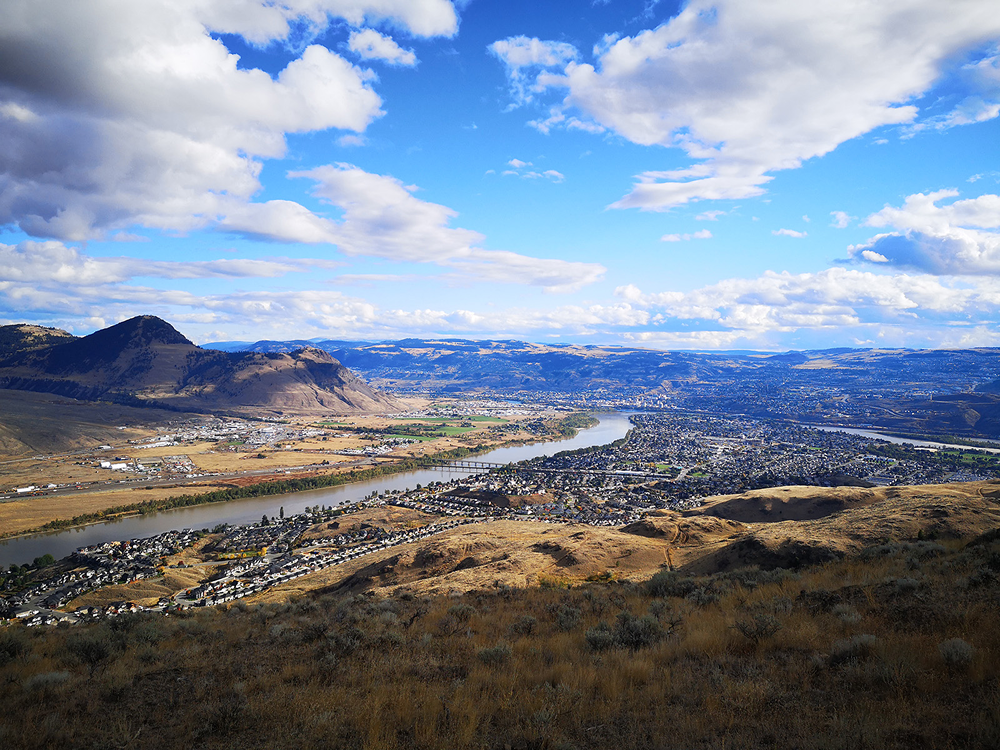 Panoramic view of the city of Kamloops, British Columbia, showing the Thompson River winding through the valley with surrounding hills, neighborhoods, and a partly cloudy blue sky.