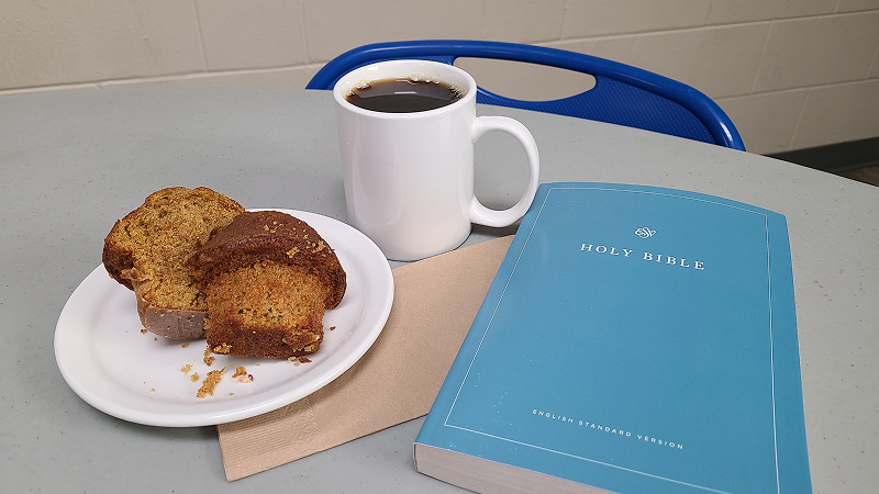 A table with a cup of coffee, a plate of sliced loaf cake, and a closed blue Holy Bible resting on a napkin, with a blue chair visible in the background.