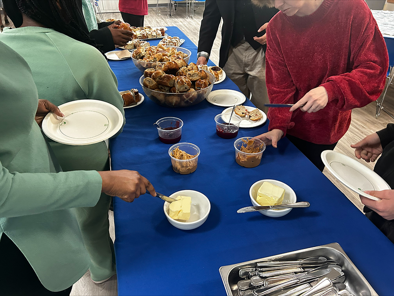 People gathered around a table covered with a blue tablecloth, serving themselves coffee, baked goods, and snacks from shared dishes and trays.