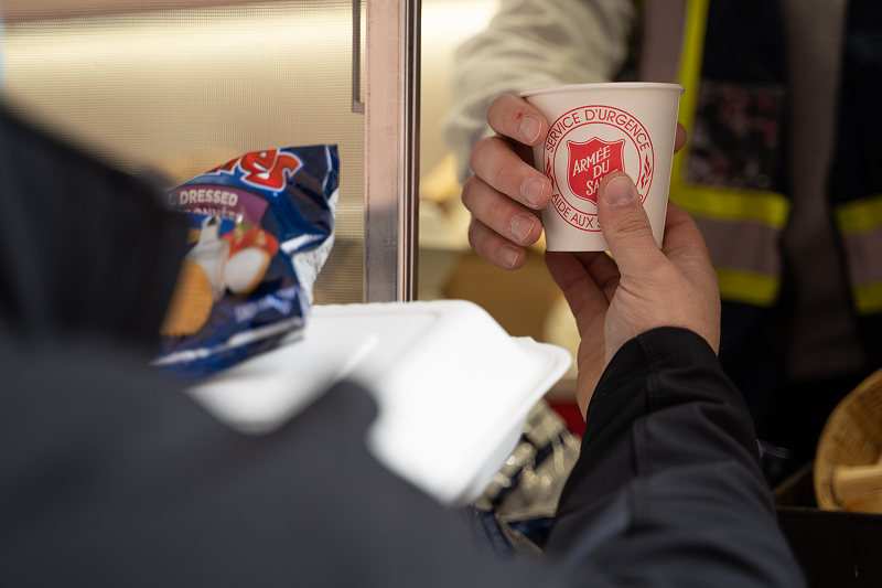 Close-up of hands holding a hot drink cup with a Salvation Army logo, with a bag of chips and a takeout container visible nearby, suggesting food and community support services.