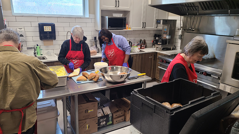 Several volunteers wearing aprons work together in a commercial kitchen, preparing food at a central counter with trays, bowls, and kitchen equipment visible around them.