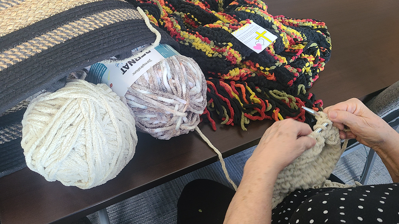 Hands working with yarn during a crafting activity at a table, with balls of colorful yarn and partially completed knitted pieces spread out nearby.
