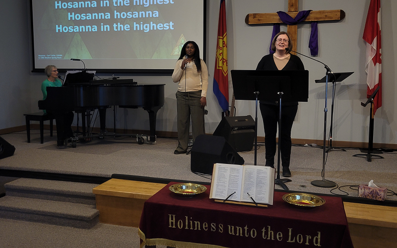 Worship service taking place inside the Kamloops Salvation Army church, showing several people singing and speaking from a stage with microphones, a piano, a wooden cross on the wall, and worship text projected above the platform.