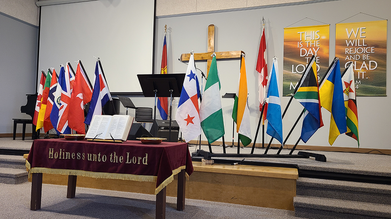 Interior of the Kamloops Salvation Army church sanctuary with a wooden cross at the center of a raised platform, surrounded by international flags, a lectern with an open book, and banners displayed on the wall behind.