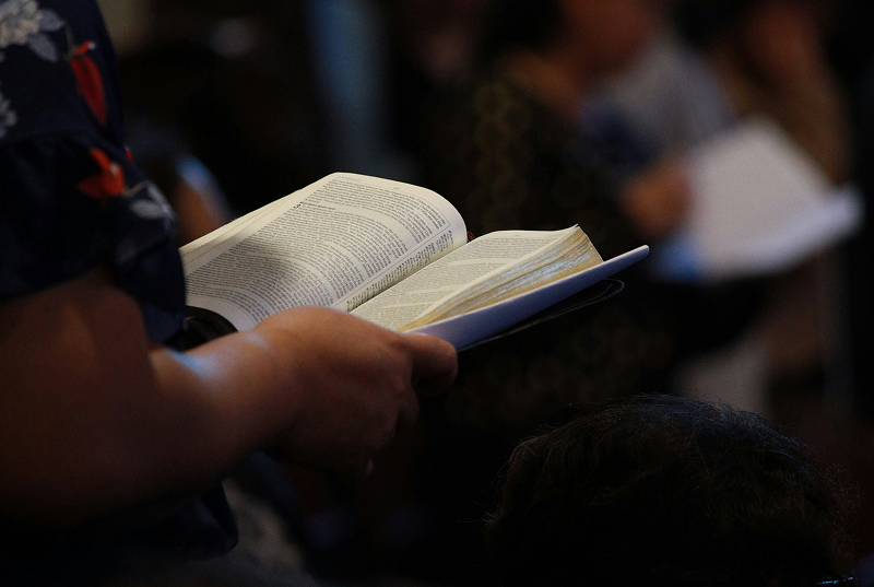 Close-up of hands holding an open Bible in a dimly lit indoor setting, with other seated people softly out of focus in the background.