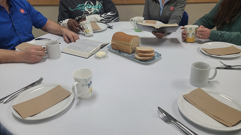 People seated around a table set with white mugs, plates, utensils, a loaf of bread on a serving board, and open books, suggesting a shared meal and discussion setting.