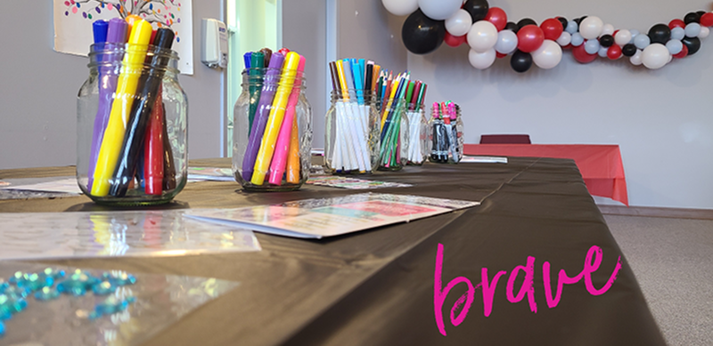 Table set up for an arts and crafts activity with jars filled with colorful markers and pens, activity sheets laid out on a tablecloth, and the word brave in the lower right corner.