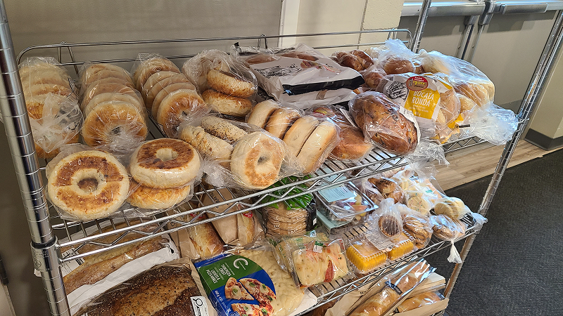 Metal shelving unit filled with assorted loaves of bread, buns, bagels, and baked goods wrapped in plastic, arranged across multiple shelves indoors.