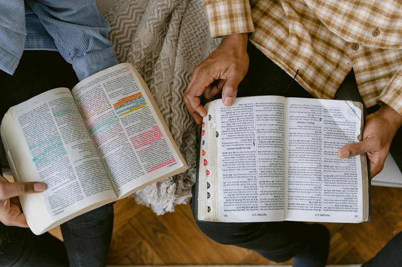 Close-up view of two open Bibles resting side by side, with hands holding and reading the pages, showing highlighted and annotated passages.
